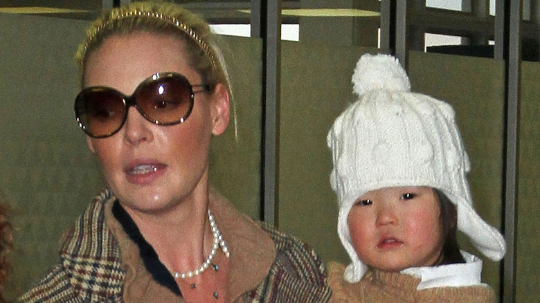 Katherine Heigl wearing a peacoat and dark sunglasses holding daughter Naleigh wearing.a white winter hat