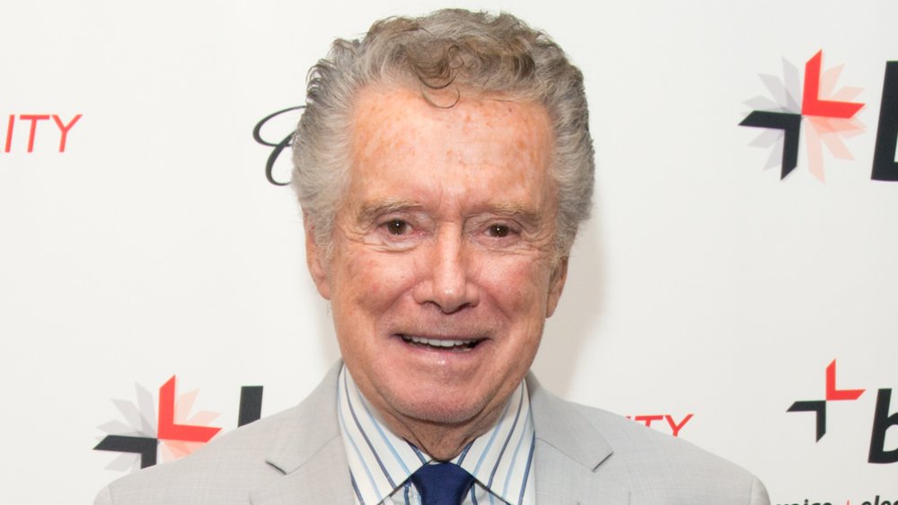 Regis Philbin in a tan suit and blue tie, with a small smile at a red carpet event