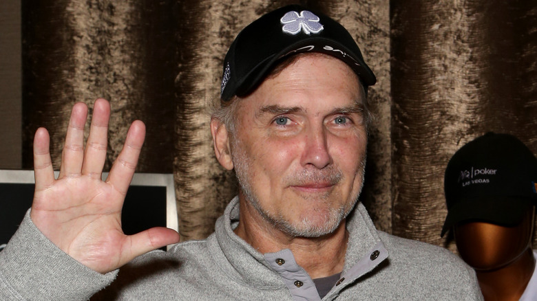 Norm Macdonald smiling and waving while wearing a black hat and grey sweatshirt