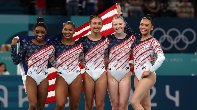 The 2024 US women's Olympic gymnastics team wearing sparkling red, white and blue leotards holding an American flag behind them