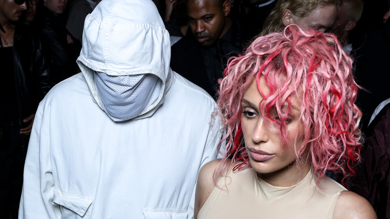 Bianca Censori with pink hair in a crowd next to Kanye West is wearing all white with his face covered