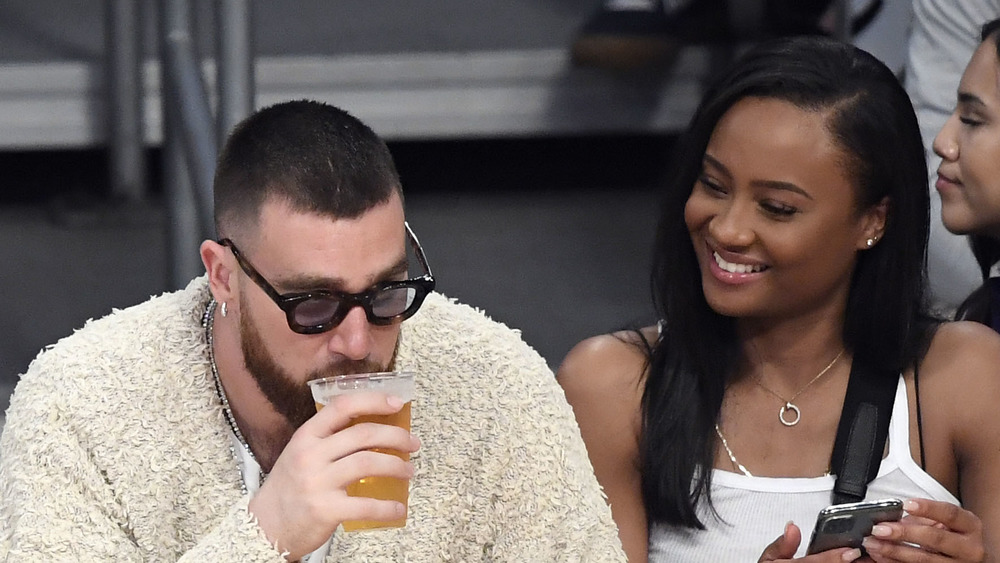 Travis Kelce, drinking a beer, court side at NBA game, wearing sunglasses; Kayla Nicole, long hair down, smiling at Travis Kelce, white tank top, court side at basketball game