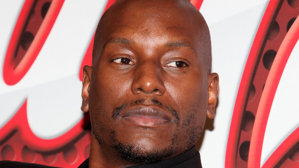 Tyrese Gibson in an all-black suit, with a serious expression, sitting back during an interview