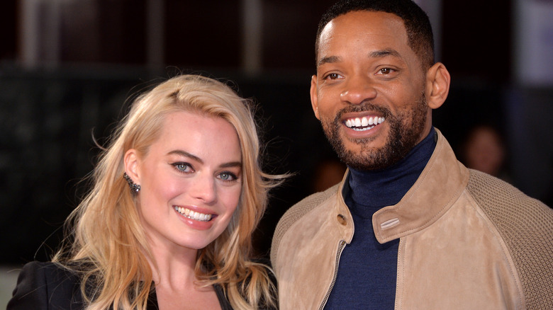 Margot Robbie in a black outfit, Will Smith in a tan bomber jacket and blue turtleneck, both smiling at a screening of Focus