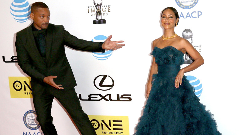 Will Smith in a black suit, dark blue shirt, and black bow tie, Jada Pinkett Smith in a blue dress and gold necklace, standing far apart on the red carpet