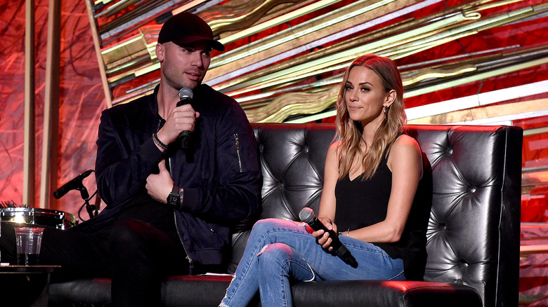 Mike Caussin, wearing dark clothes and a hat, speaking into a microphone;  Jana Kramer, sitting on a couch, looking annoyed, wearing jeans, 2019 photo