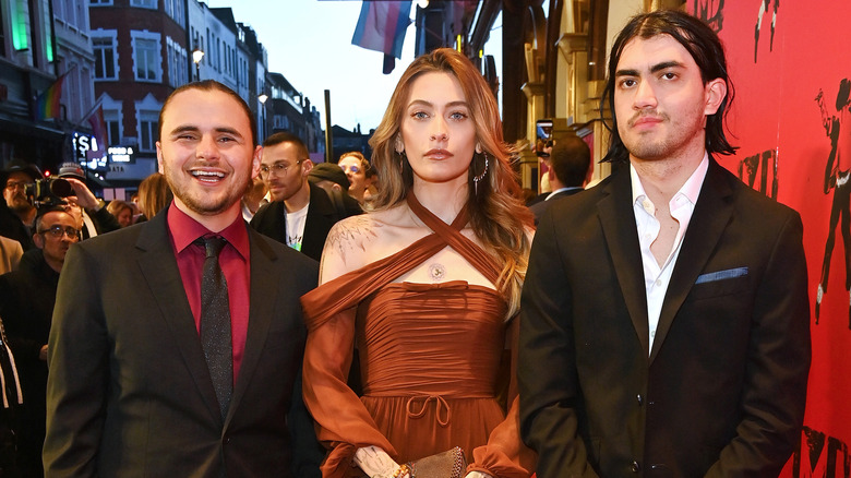 Prince Jackson wearing a black jacket, red shirt, and black tie; with Paris Jackson wearing a rust-colored criss-crossed dress; with Bigi Jackson wearing a black jacket and white shirt