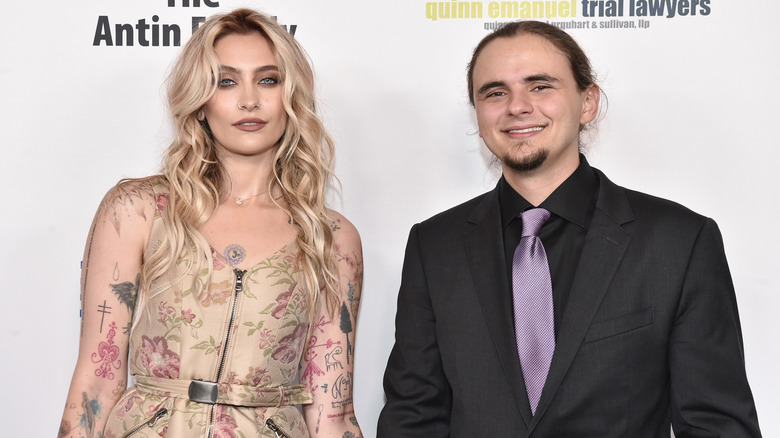 Paris Jackson wearing a tan floral dress with Prince Jackson wearing a black jacket, black shirt, and purple tie