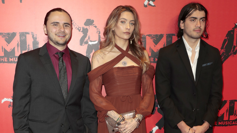 Prince Jackson wearing a black jacket, red shirt, and black tie; with Paris Jackson wearing a rust-colored criss-crossed dress; with Bigi Jackson wearing a black jacket and white shirt