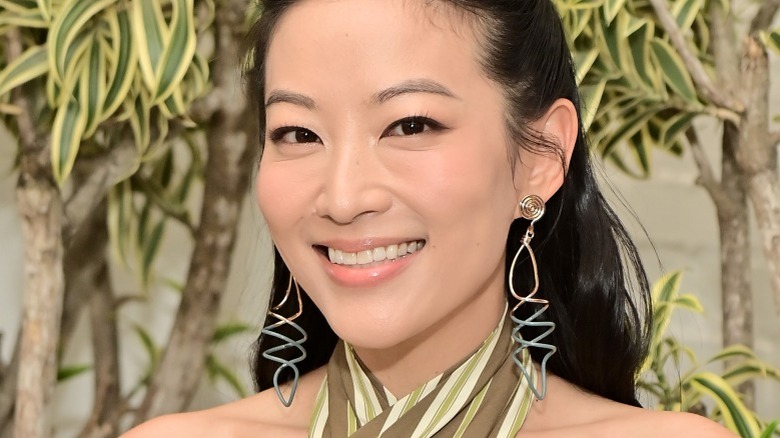 Arden Cho smiling big earrings in front of trees