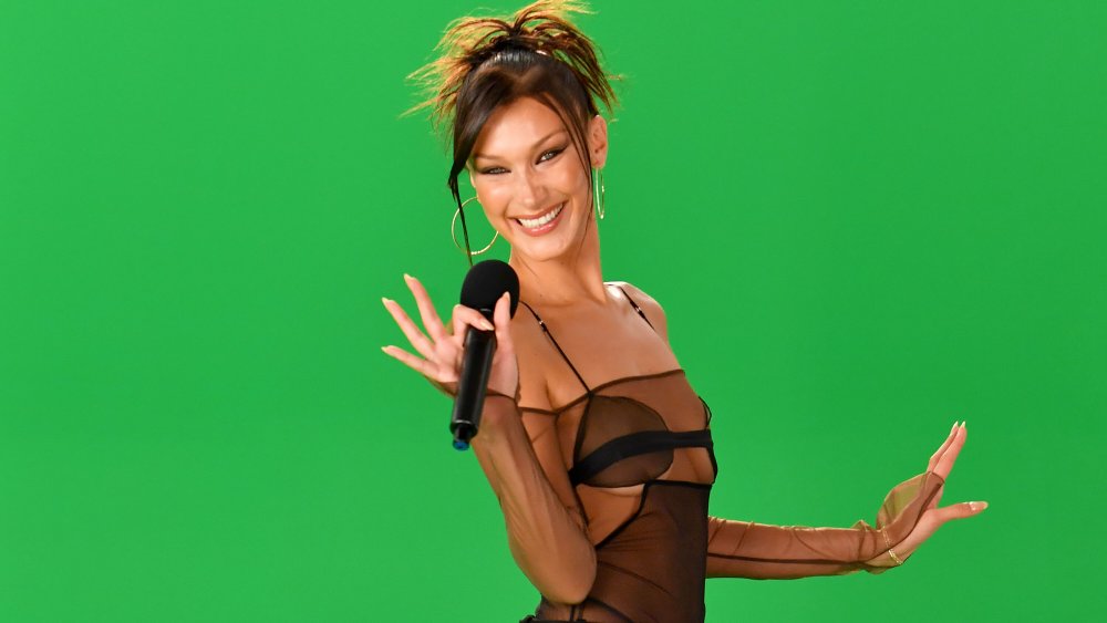 Bella Hadid in front of green screen, hair up and holding microphone