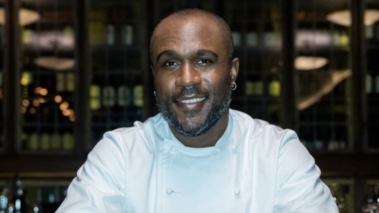 Chef Cliff Crooks, smiling, sitting at a table in a restaurant, wearing a white chef's outfit