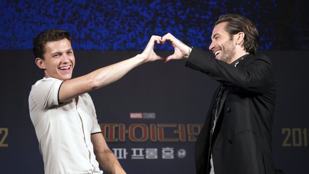 Tom Holland and Jake Gyllenhaal laughing while forming a heart with their hands at a Spider-Man: Far From Home press event