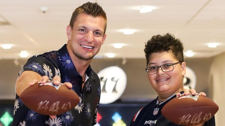 Rob Gronkowski smiling holding a football with a child wearing glasses and a Patriots football jersey