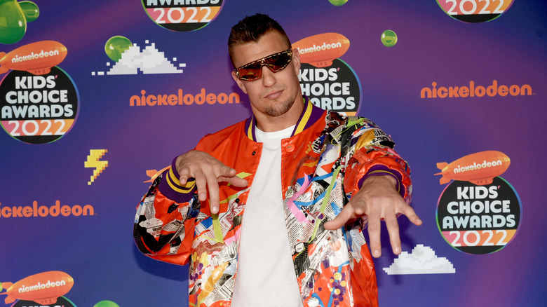 Rob Gronkowski wearing an orange and neon-patterned jacket, a white t-shirt, and dark sunglasses while pointing at the camera