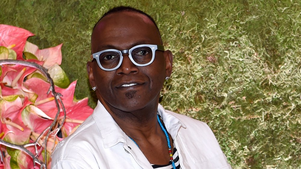 Randy Jackson with thick-framed blue glasses standing in front of flowers