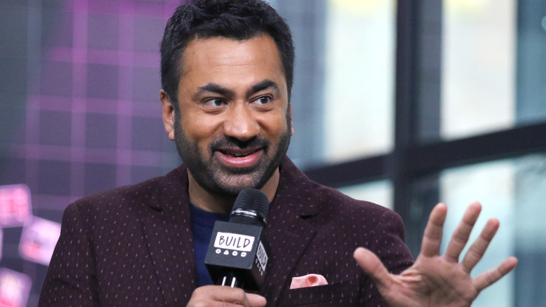 Kal Penn smiling and talking animatedly into a microphone