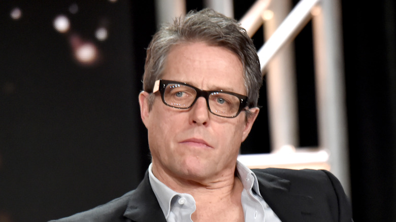 Hugh Grant sits on stage while wearing glasses