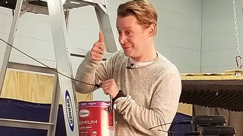 Macaulay Culkin carrying a can of paint up a ladder while smiling and giving a thumb's up