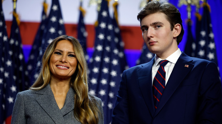 Melania Trump smiling next to Barron Trump in front of several American flags