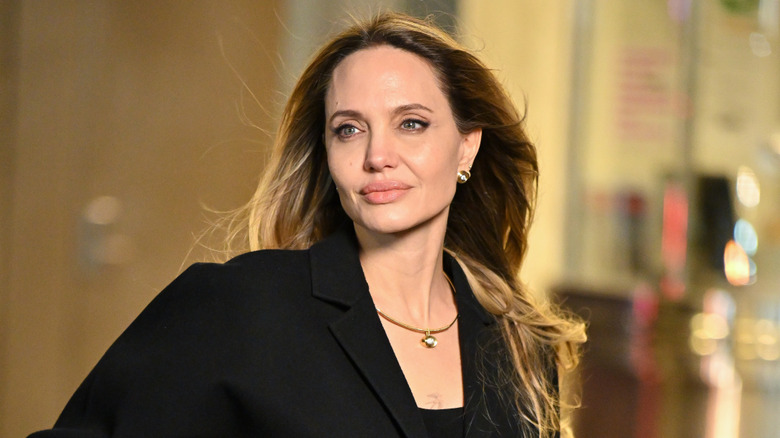 Angelina Jolie wearing a black blazer and gold jewelry and turning as wind blows her hair